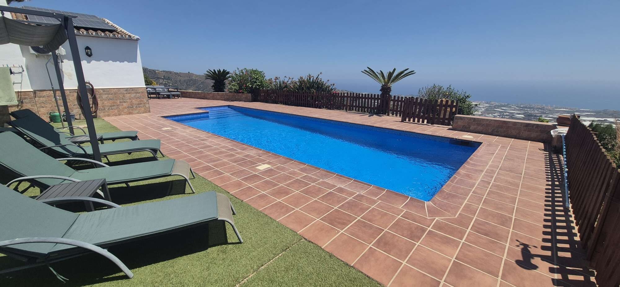Une piscine rectangulaire dotée d’une terrasse carrelée, entourée d’une clôture en bois, et offrant une vue sur un paysage côtier.