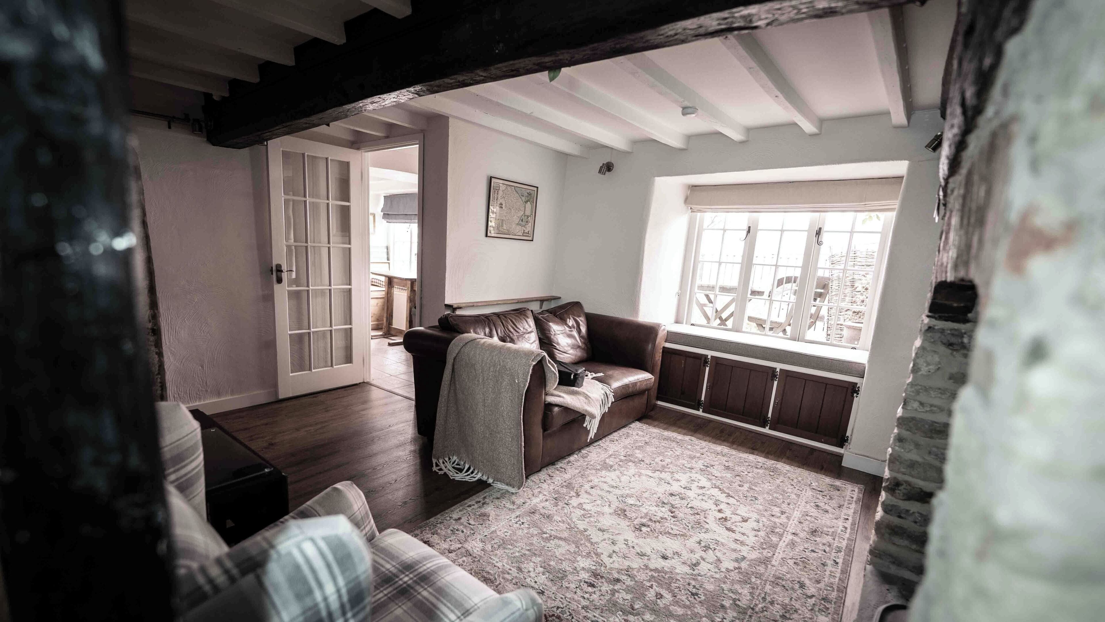 Una sala de estar acogedora con un sofá de cuero marrón, un sillón estampado y un tapiz con diseño. Un ventanal grande proporciona luz natural, y una puerta conduce a otra habitación.