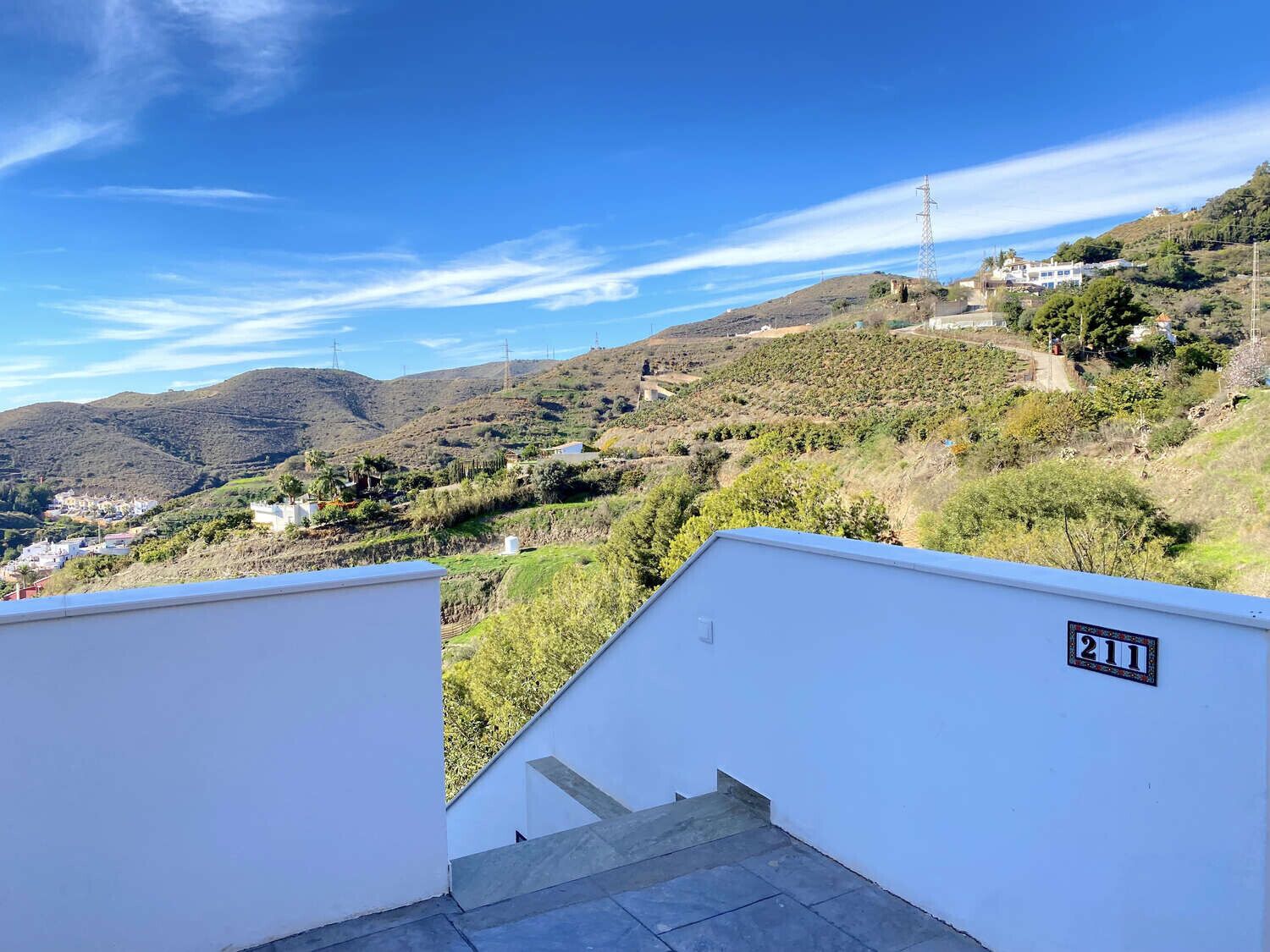 Una scala conduce a un balcone con vista su un paesaggio collinare, con edifici e linee elettriche.