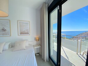 A hotel room with a bed, a nightstand, a lamp, and a large window with a view of the sea and cityscape. - Balcon del Mar Seaview 211 by Casasol (Nerja)