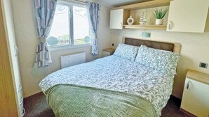 Een slaapkamer met een bed, een raam met gordijnen, een radiator, een nachtkastje, een plank met decoratieve objecten en een klein kastje.