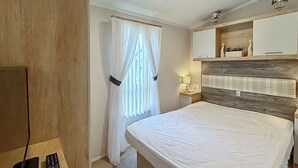 A hotel room with a bed, a nightstand, a lamp, a TV, and a window with curtains. - Swift Bordeaux Escape - UKC9640 (Towyn)