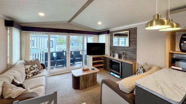 A modern living room with a sofa, a dining table, a kitchen area with a TV, and a sliding glass door leading to an outdoor patio. - Swift Bordeaux Escape - UKC9640 (Towyn)