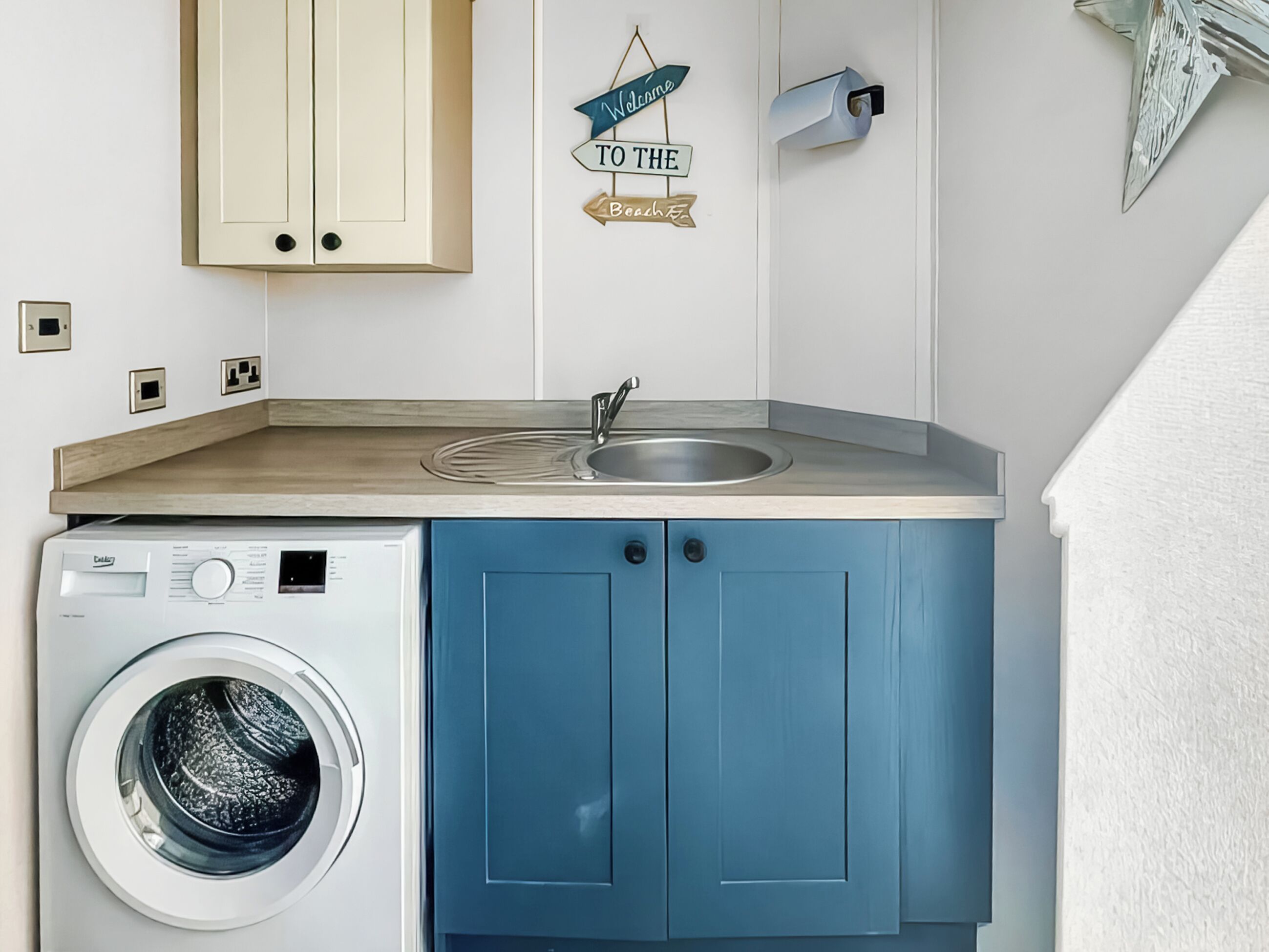 A compact kitchen with a washing machine, a sink, and a decorative welcome sign.