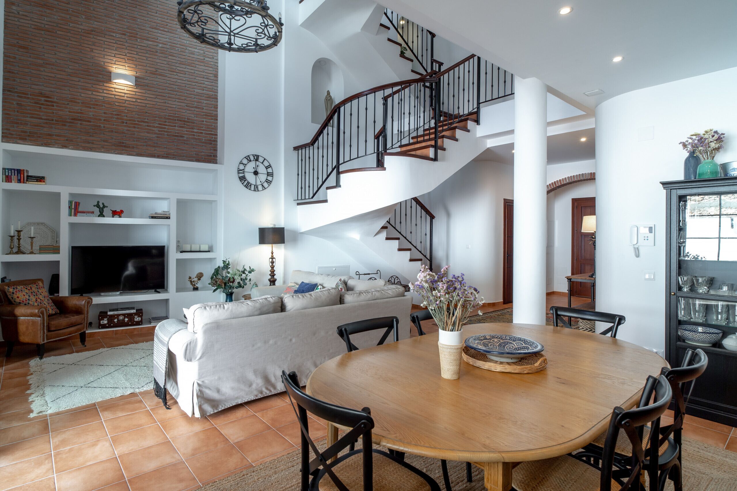A spacious living area with a wooden dining table, a sofa, and a staircase leading to an upper level.