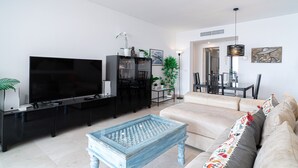 A modern living room with a flat-screen TV, a black entertainment unit, a light blue coffee table, a beige sofa, and a dining area with chairs and a table. - Calaceite 5101 Ocean Paradise by Casasol (Torrox)