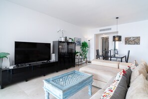 Ein modernes Wohnzimmer mit einem Flachbildfernseher, einer schwarzen Unterhaltungsanlage, einem hellblauen Couchtisch, einem beigefarbenen Sofa und einem Essbereich mit Stühlen und einem Tisch.