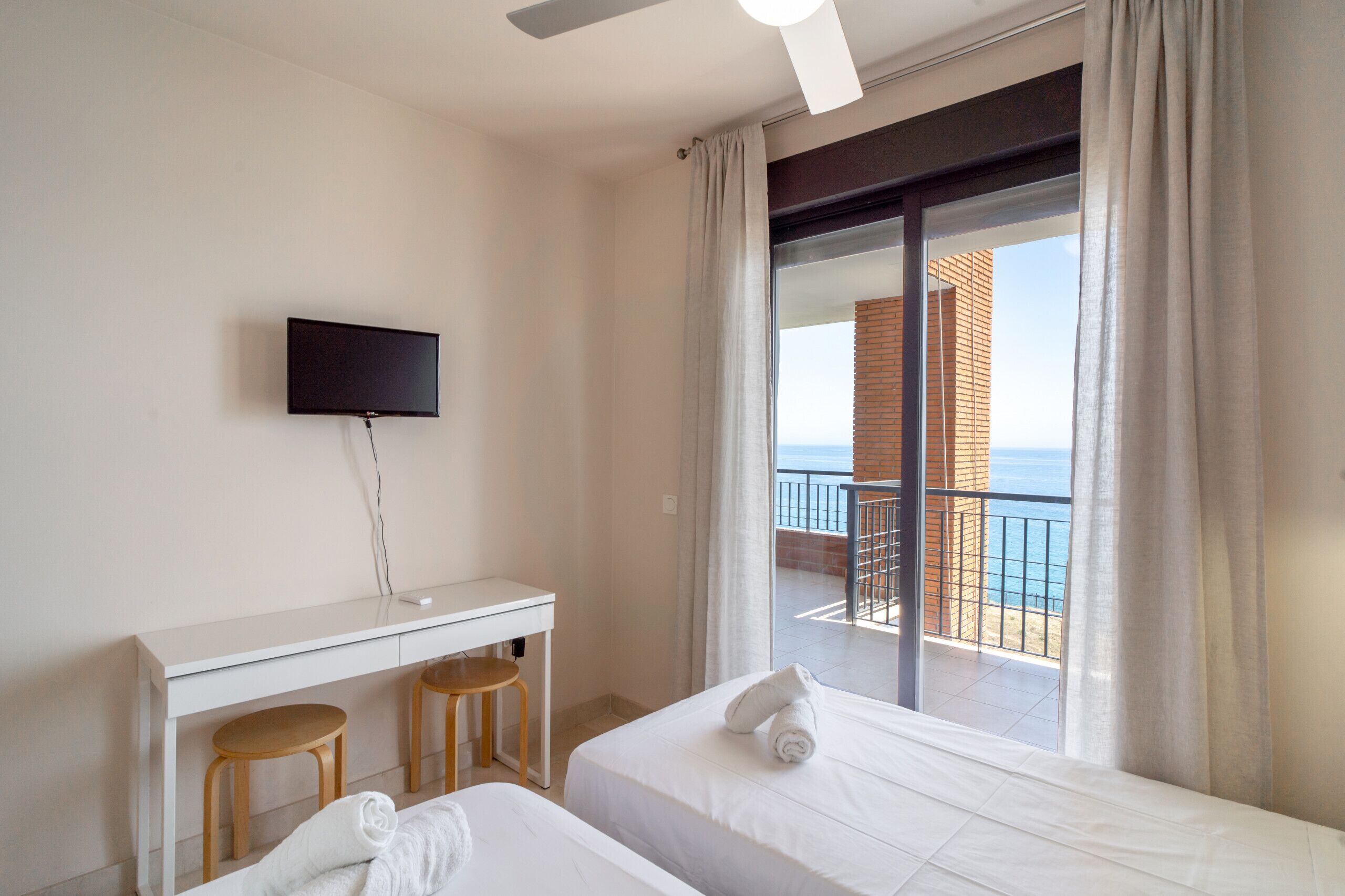 Una habitación de hotel con balcón, un televisor montado en la pared y dos taburetes de madera.