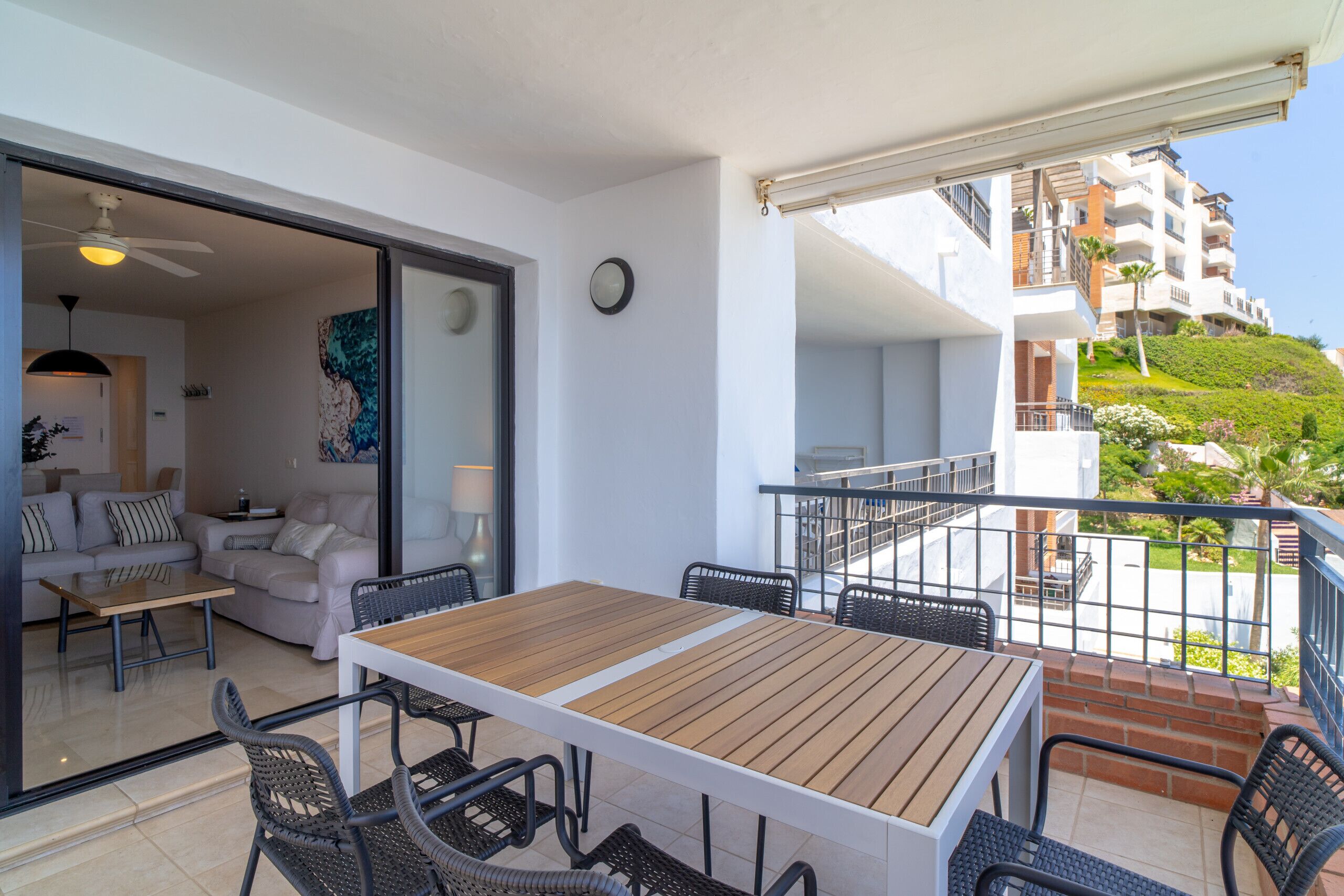Ein Balkon mit Holztisch und -stühlen, einer Sitzecke und Blick auf eine Hügellandschaft mit Gebäuden.