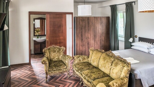 A hotel room with a bed, two ornate armchairs, a wooden wardrobe, a mirror, and a modern pendant light.