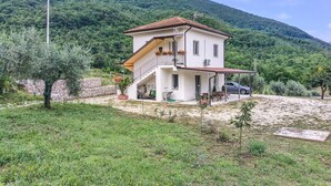 A two-story house with a balcony, surrounded by greenery and a mountain in the background.