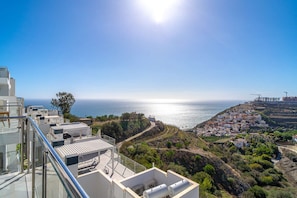 Ein Küstenblick mit klarem Himmel, ein Gebäude mit Glasbalkon mit Blick auf das Meer und eine Hügelkuppe mit Wohngebäuden.