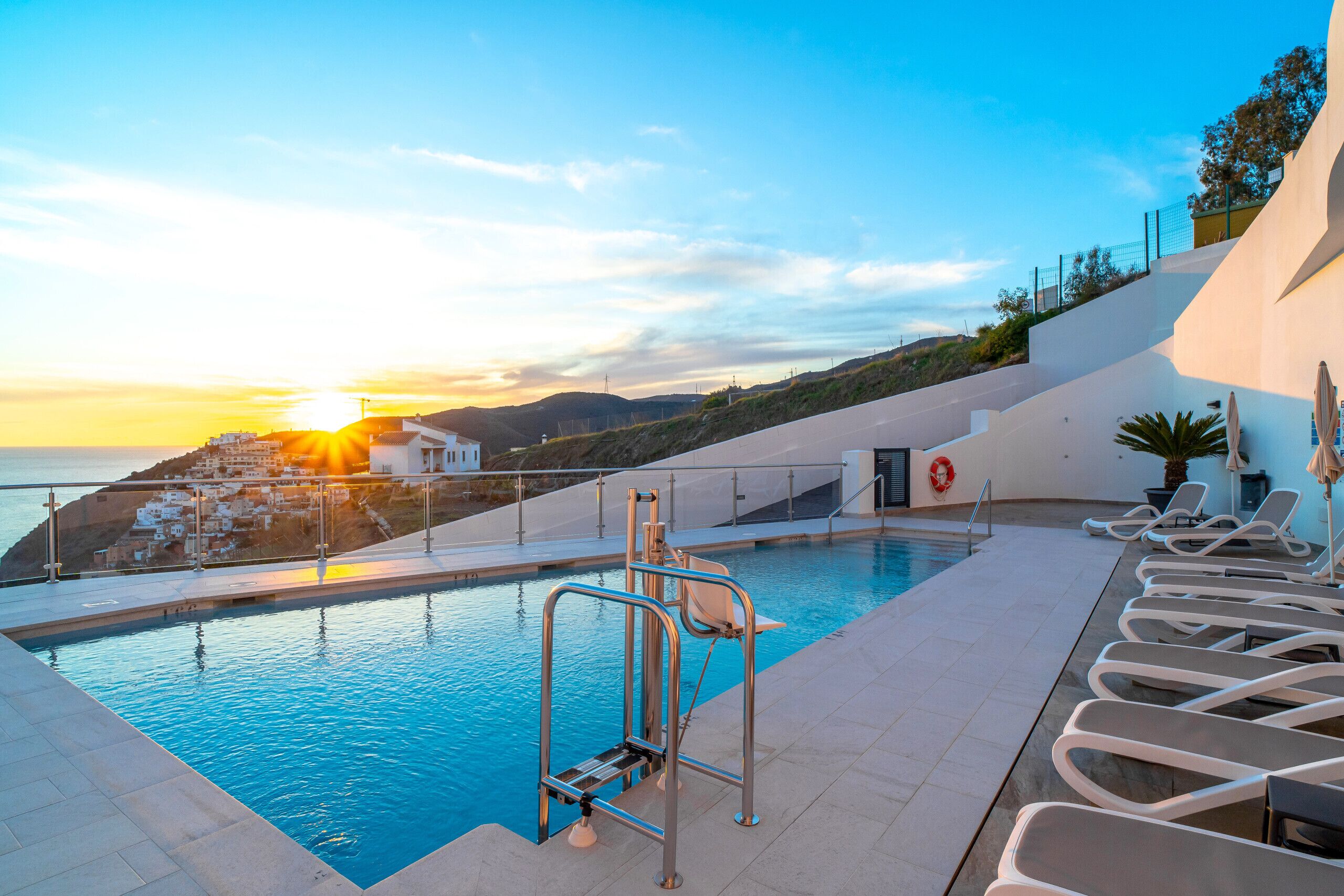 Une piscine dotée de chaises longues et d’une bouée de sauvetage, offrant une vue sur la côte au coucher du soleil.