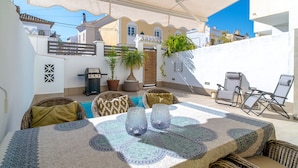 Eine Terrasse mit Tisch für zwei Personen, Korbstühle, einem Grill und Kübelpflanzen.