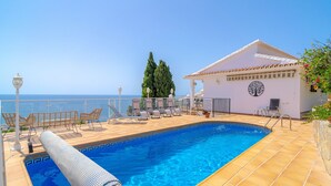 Area piscina con vista libera sul mare, sedie a sdraio e un edificio con emblema circolare.