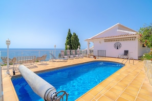 Ein Poolbereich mit freiem Blick auf das Meer, Liegestühle und ein Gebäude mit kreisförmigem Wappen.