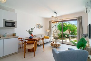 Ein modernes Wohnzimmer mit Sofa, Esstisch und Stühlen. Es gibt eine Küchenzeile mit Mikrowelle und Schränken. Ein großes Fenster bietet Blick auf Grünflächen außerhalb.