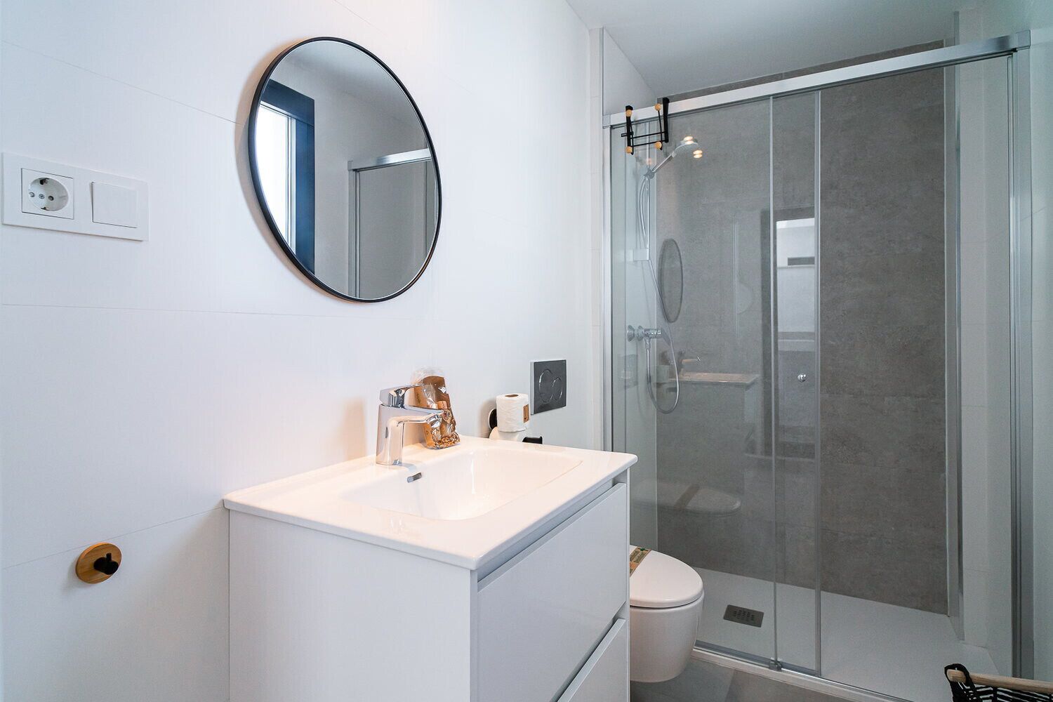 Ein modernes Badezimmer mit einem wei ßen Waschtisch, einem runden Spiegel, einer Duschkabine aus Glas und einer Toilette.