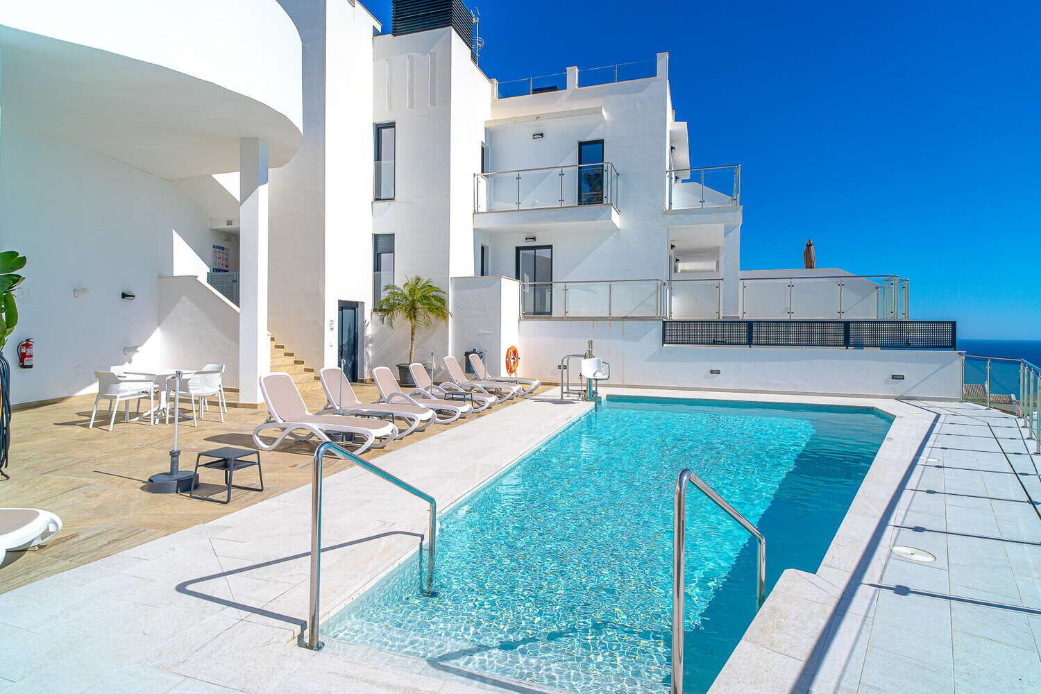 Un área de piscina moderna con piscina azul cristalina, sillones reclinables blancos y una escalera que conduce a un edificio con balcones.