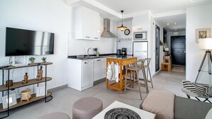 A modern kitchen with white cabinets, a central island with bar stools, a flat-screen TV, and a dining area with a sofa and ottomans. - Terrazas de Ladera A2 Luxury by Casasol (Nerja)
