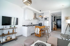 Eine moderne Küche mit weißen Schränken, einer zentralen Kochinsel mit Barhocker, einem Flachbildfernseher und einem Essbereich mit einer Couch und Poufs.