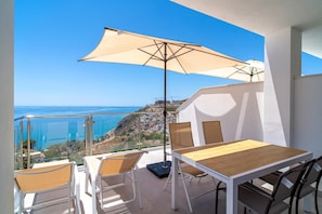 Ein Balkon mit Tisch und Stühlen, einem großen Sonnenschirm und Blick auf die Küste.