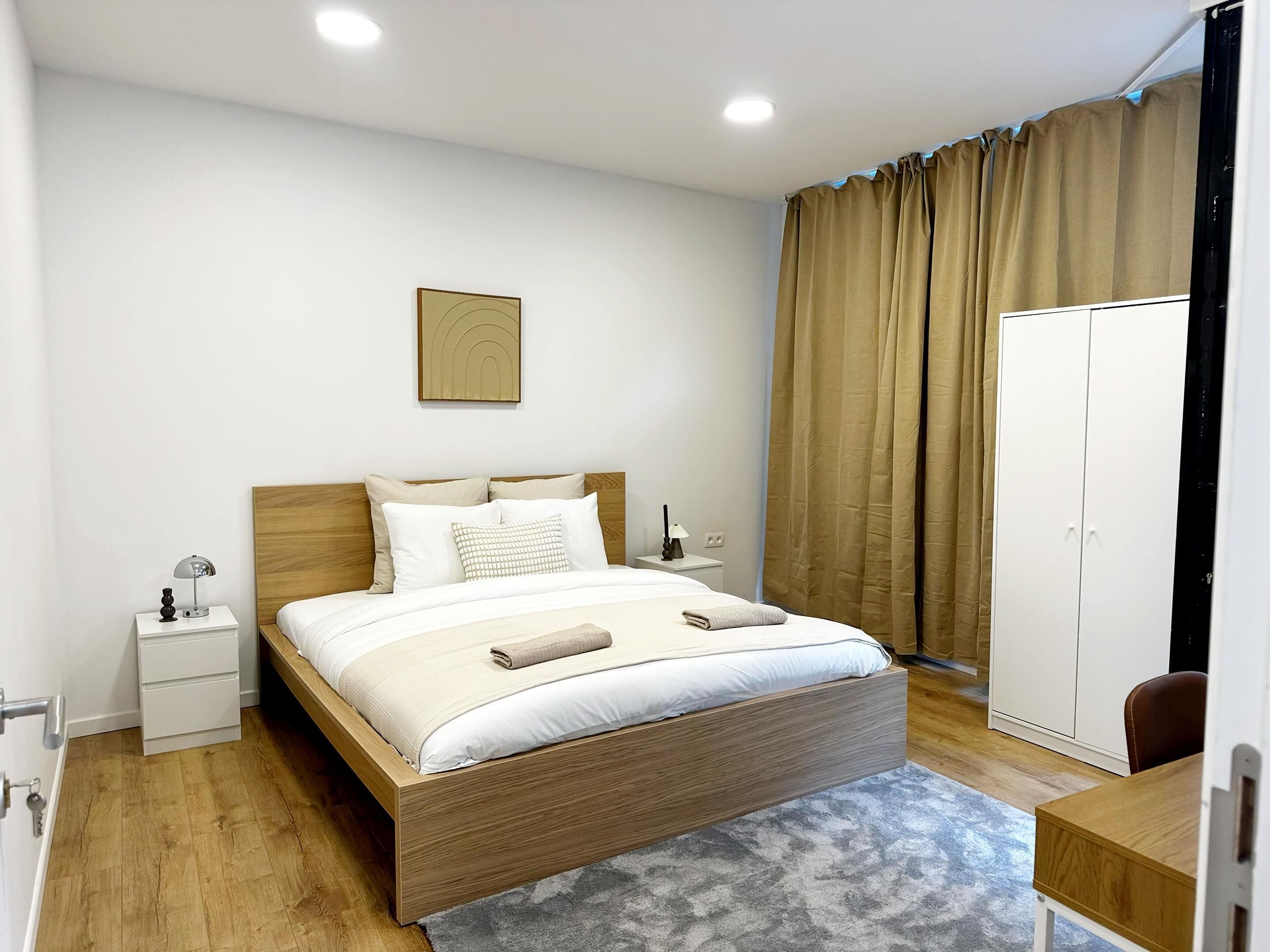 Ein modernes Schlafzimmer mit einem hölzernen Bett, weißer Bettwäsche, einem Nachttisch mit Lampe, einem Kleiderschrank und einem gerahmten Bild an der Wand.