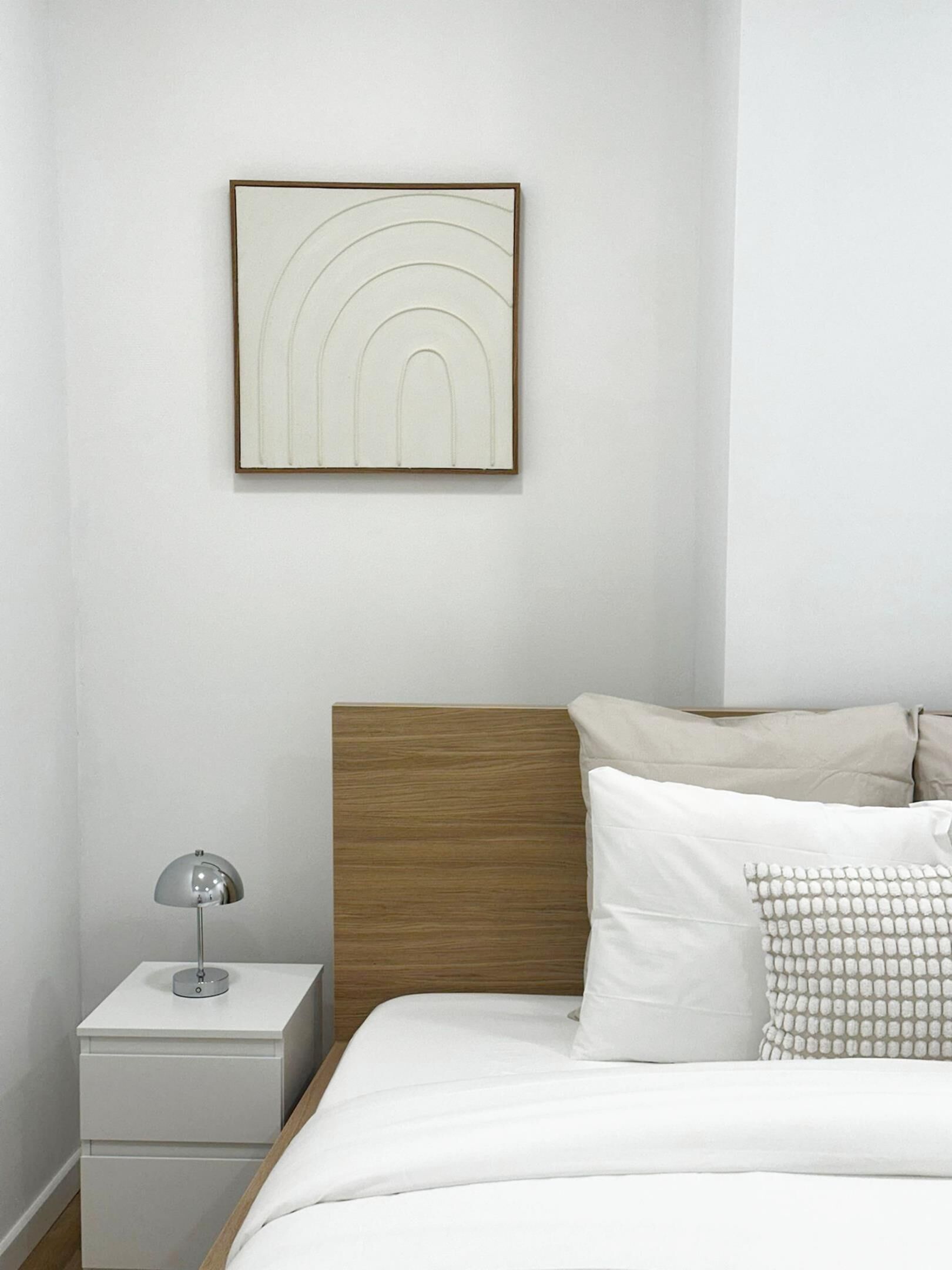 Een netjes ingerichte slaapkamer met een houten hoofdbord, een nachtkastje met een lamp, en een lijst met abstracte kunst aan de muur.