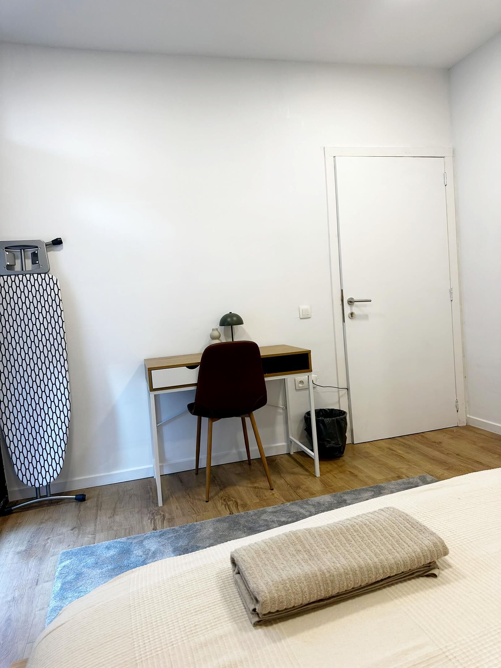 A bedroom with a bed, a desk, a chair, a door, and an ironing board.