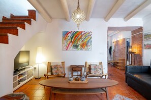 Ein Wohnzimmer mit einem Holztisch, zwei Korbstühlen, einem schwarzen Ledersofa, einem Fernseher und einem Bild an der Wand.