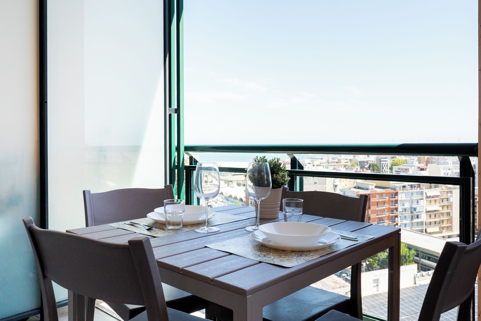 Et spisebord med stole, en potteplante og vinglas på en balkon med udsigt over byen.