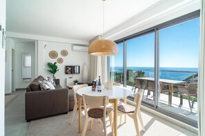 Ein modernes Wohnzimmer mit einer Couch, einem Esstisch, Stühlen, einem Fernseher und einem großen Fenster mit Meerblick.