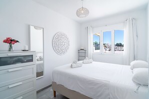 Ein helles Schlafzimmer mit Bett, Kommode, Spiegel und Fenster mit Blick auf Gebäude.