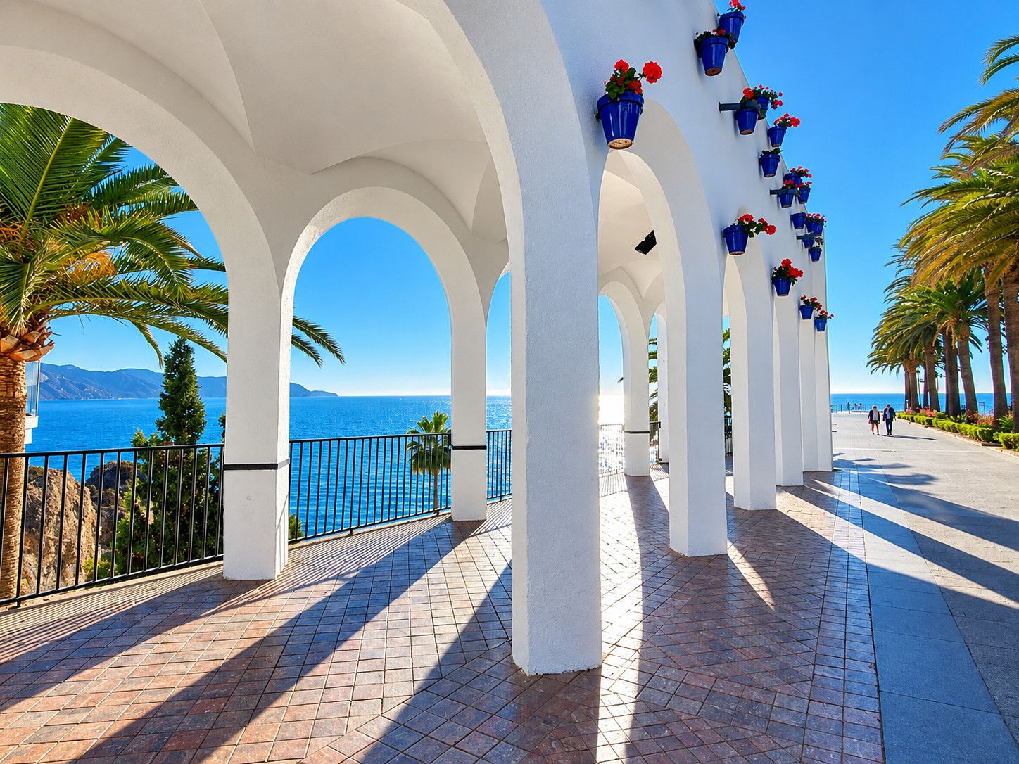 A coastal promenade with white arches, a tiled walkway, and blue flower planters.