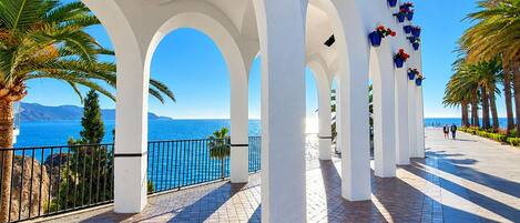 A coastal promenade with white arches, a tiled walkway, and blue flower planters.