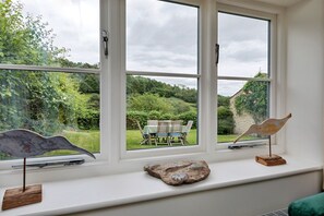 A room with a large window overlooking a garden, a rustic wooden table, and two bird-shaped sculptures on the windowsill. - The Burrows (Wass)