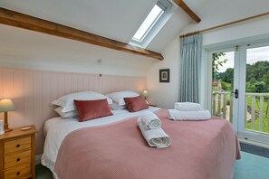 Ein Schlafzimmer mit Bett, Kissen, Nachttisch, Lampe, Fenster und Blick ins Freie.