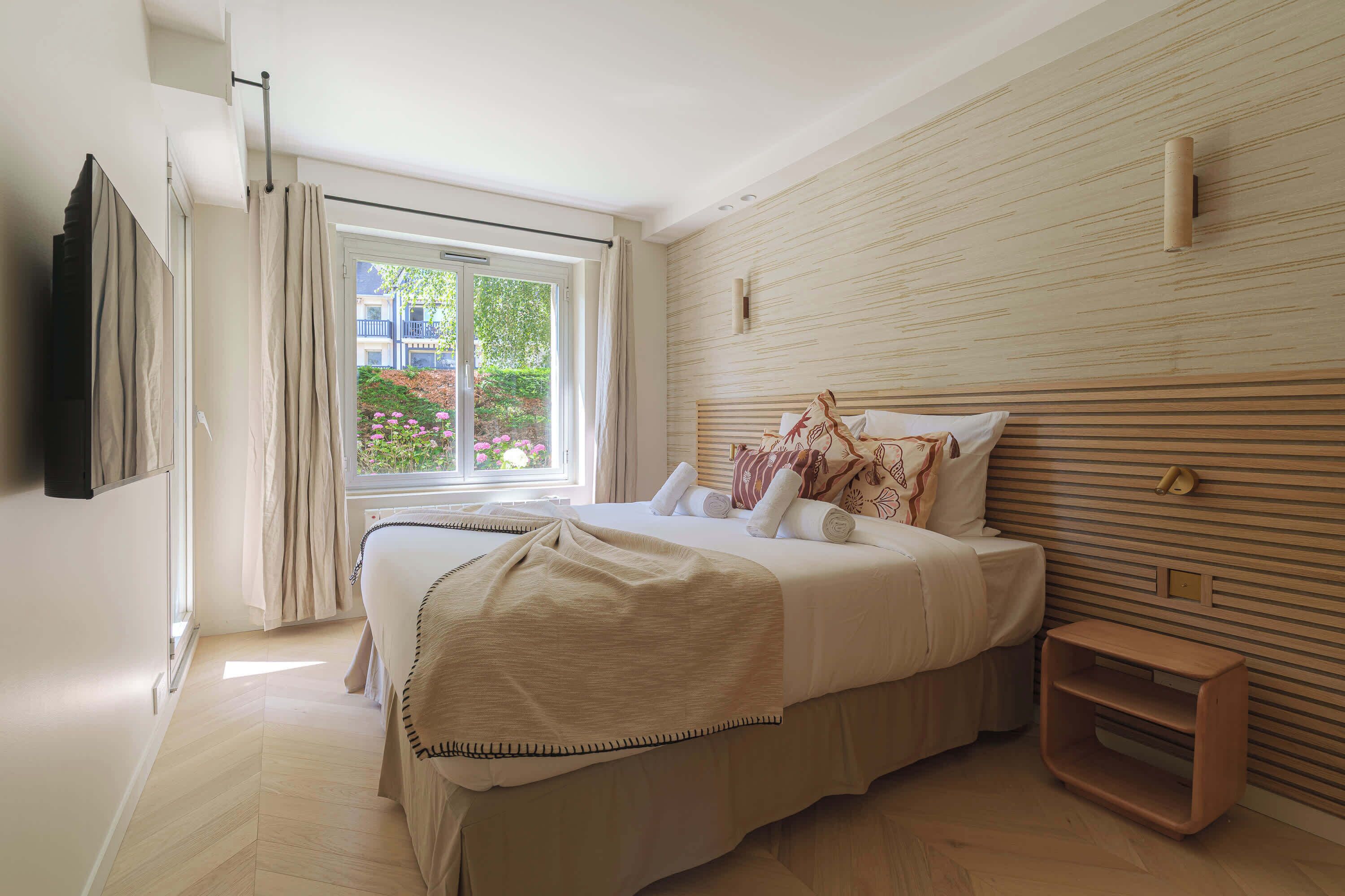 A bedroom with a bed, a bedside table, a window with curtains, and a wall-mounted light fixture.