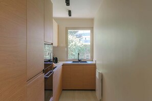 A compact kitchen with wooden cabinets, a built-in oven, and a window with a view of a building and trees. - Apartment & Private Garden - Deauville-Sévigné (Deauville)