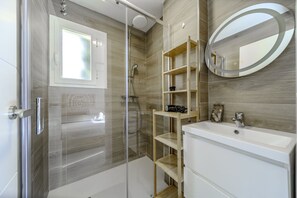 A modern bathroom with a glass-enclosed shower, a wooden shelf unit, a white sink, and a round mirror. - Jardines de Burriana Deluxe by Casasol (Nerja)