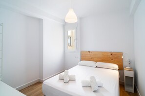 Ein schlicht eingerichtetes Hotelzimmer mit einem Holzkopfteil, einem Einzelbett mit weißer Bettwäsche, einem Nachttisch mit Lampe und Blick durch ein Fenster.