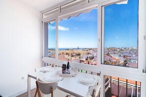 Ein Essbereich mit Tisch für zwei Personen, großen Fenstern mit Blick auf die Stadt und einem Balkon mit Geländer.
