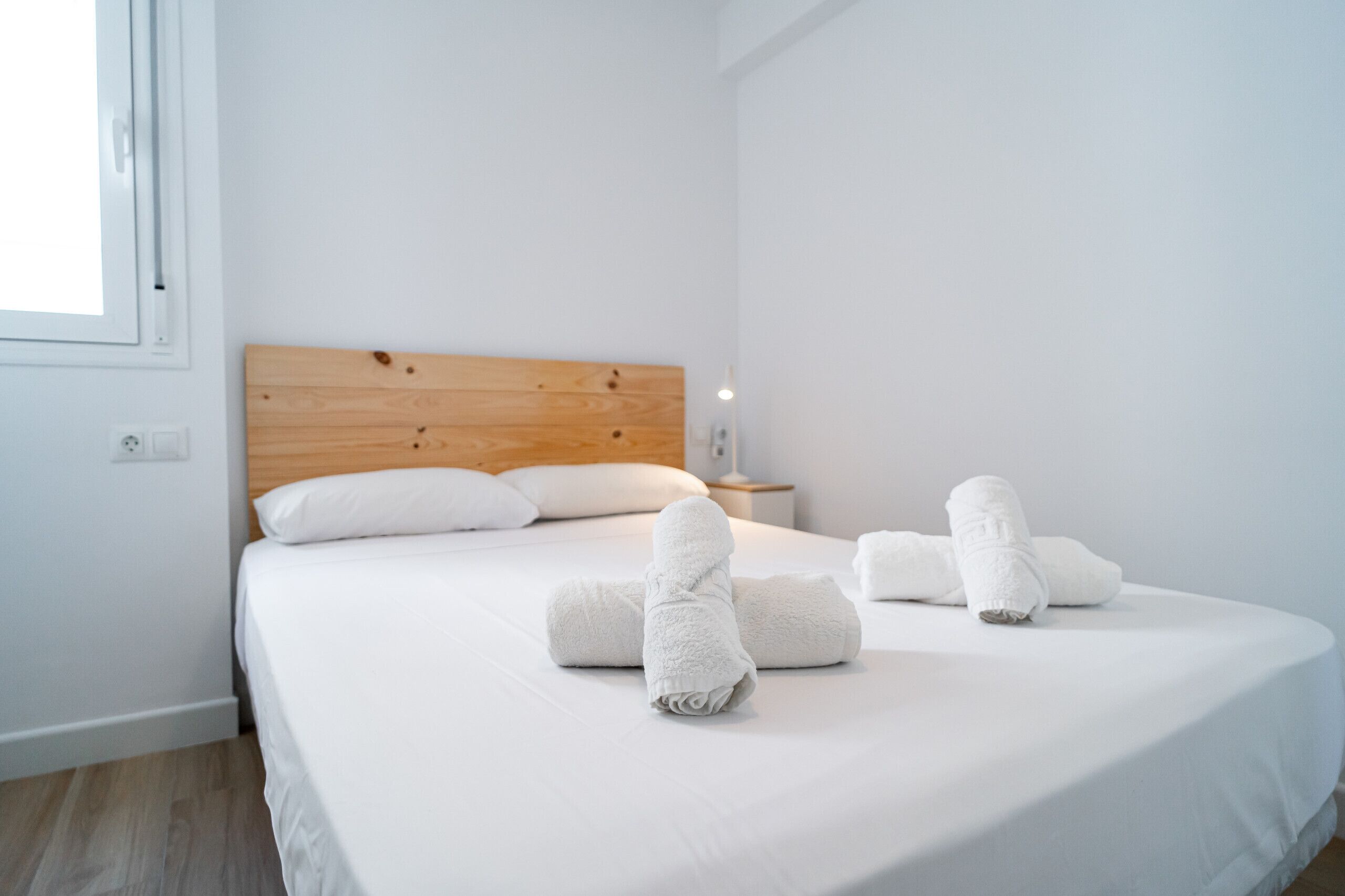 Cama bem arrumada com lençóis e toalhas brancas, cabeceira de madeira, mesa de cabeceira com abajur e janela com persianas.