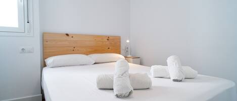 Cama bem arrumada com lençóis e toalhas brancas, cabeceira de madeira, mesa de cabeceira com abajur e janela com persianas.