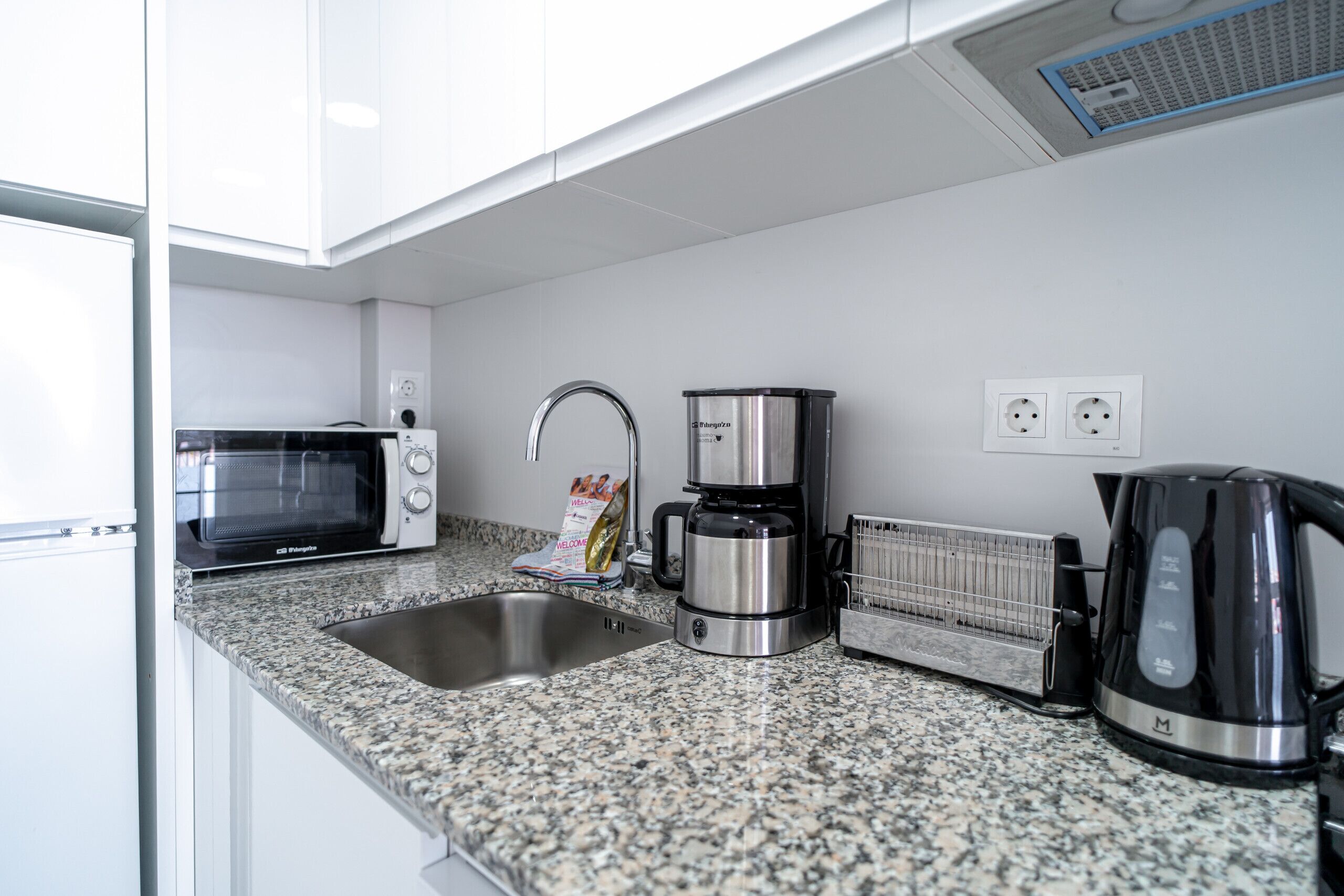 A kitchen with a microwave, coffee maker, toaster, kettle, and a sink.