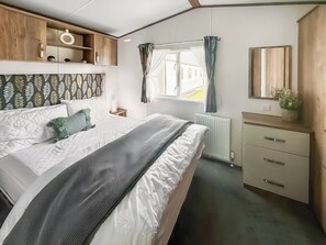 Ein Schlafzimmer mit Bett, Kommode, Spiegel, Fenster mit Vorhängen und Heizkörper.