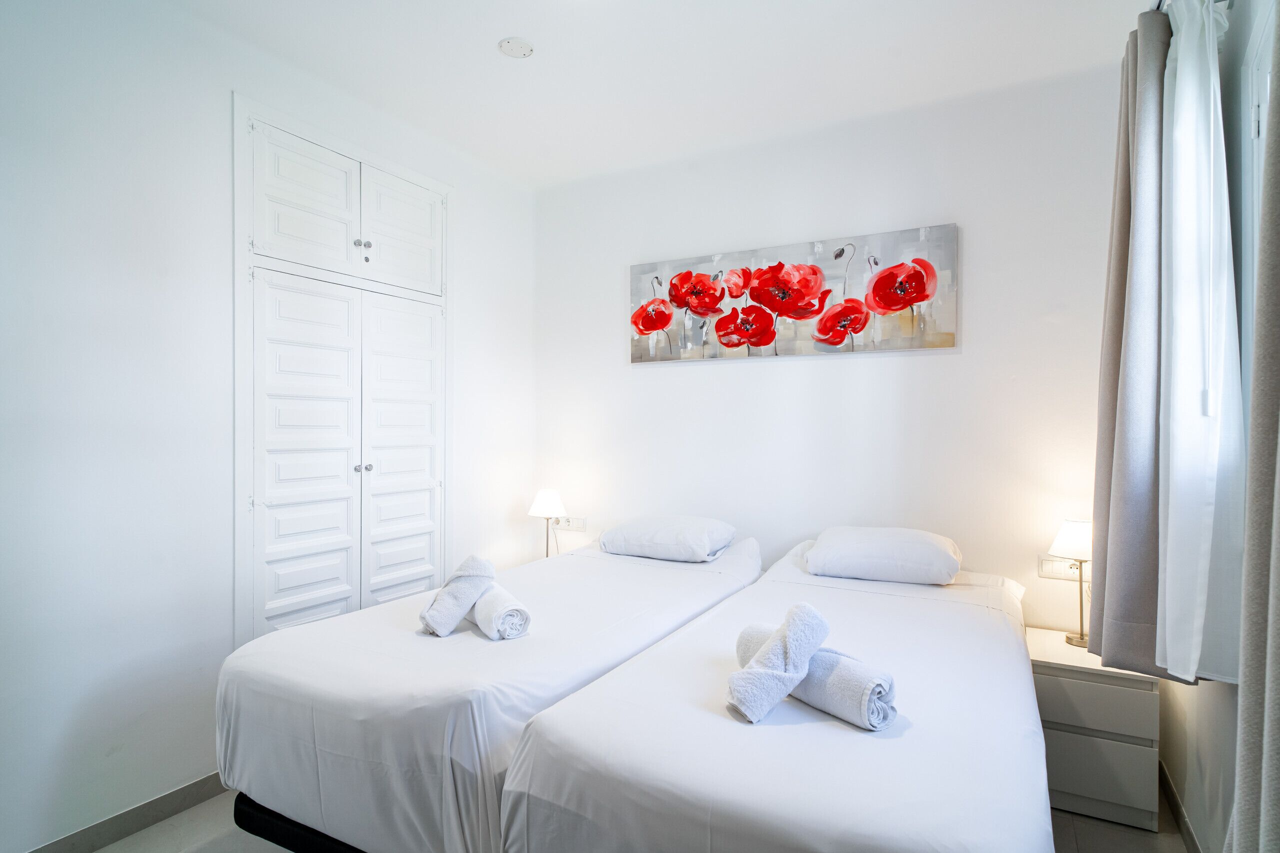 A hotel room with two beds, white bedding, a painting of red flowers on the wall, and a window with curtains.
