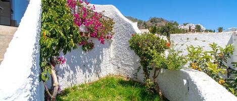 A white-walled building with a small garden area, a balcony, and a view of the surrounding landscape.
