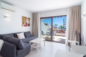 Ein modernes Wohnzimmer mit einem grauen Sofa, einem weißen Couchtisch und einem Fernsehschrank. Ein großes Schiebeglasfenster führt auf eine Terrasse mit Tisch und Stühlen.