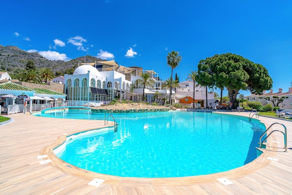 Ein großer, klarer Swimmingpool mit umlaufender Holzterrasse, vor einer Kulisse aus Gebäuden und Palmen.